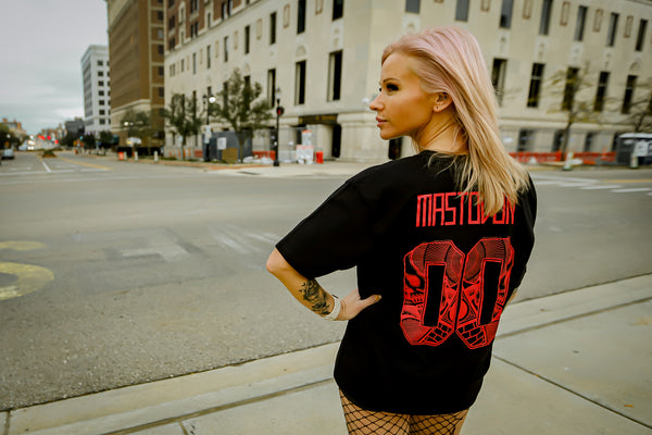 A woman with pink hair stands on a city street corner wearing a black oversized T-shirt with a large red 'Mastrobu 00' graphic on the back, paired with fishnet tights.
